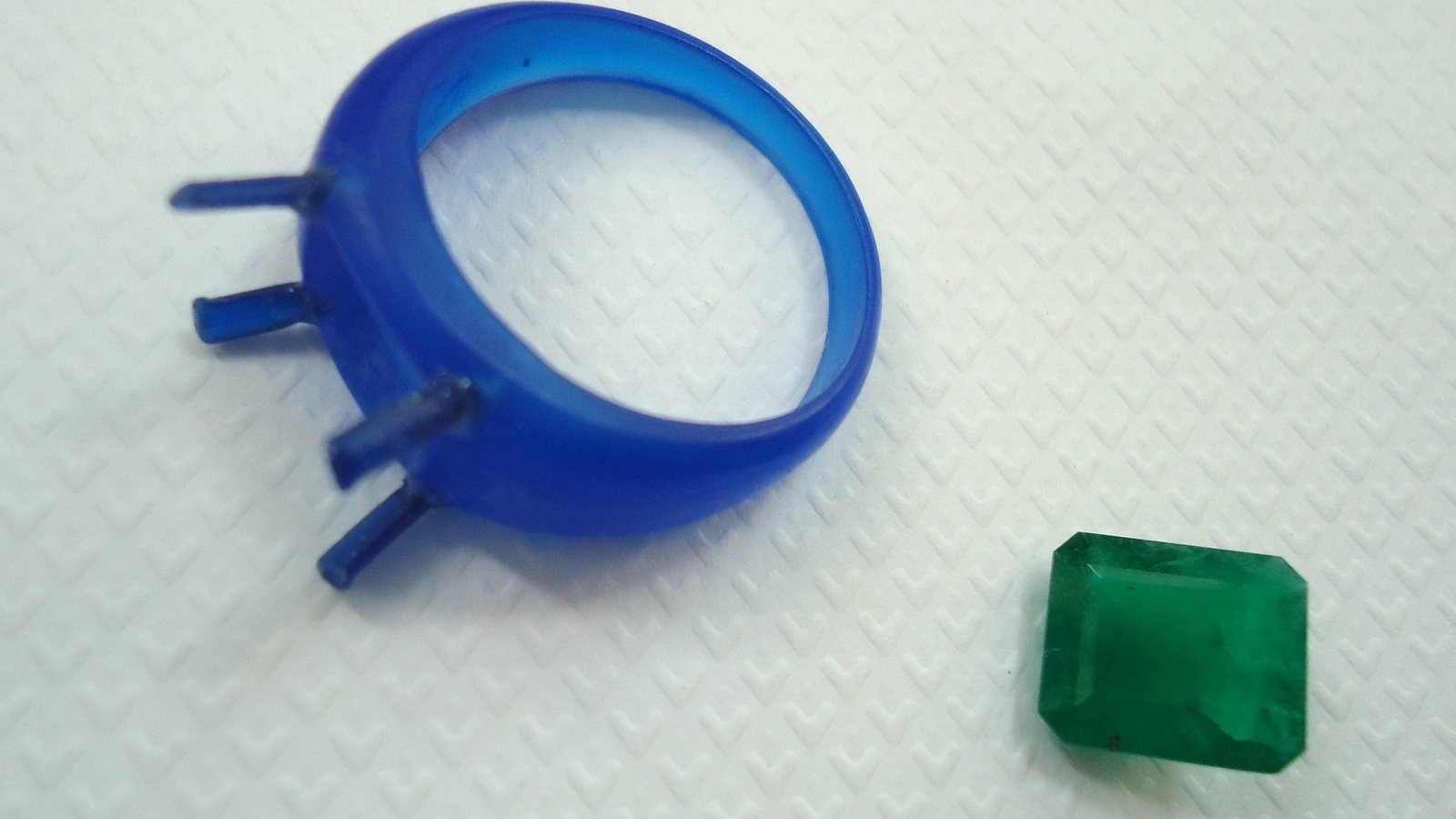Modèle de bague en cire bleue avec quatre griffes, posé à côté d'une émeraude taillée qu'il doit recevoir