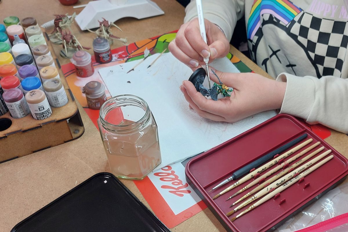 Mains qui peignent une miniature au pinceau fin, entourées de pots de peinture