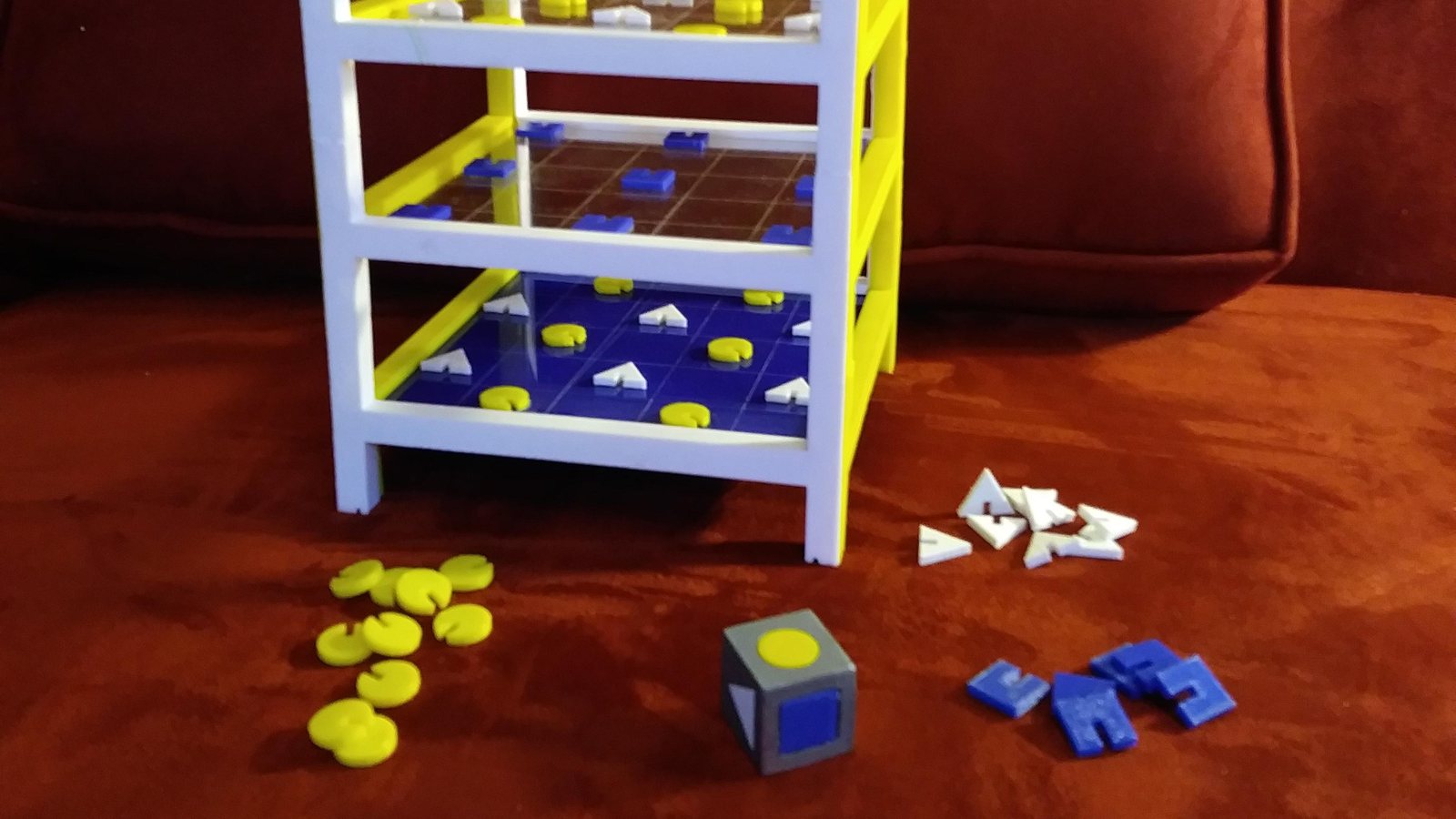 Prototype de jeu de société imprimé en 3D : tour à étages en PLA blanc et jaune, jetons jaunes, pions triangulaires blancs, pièces bleues et dé imprimé