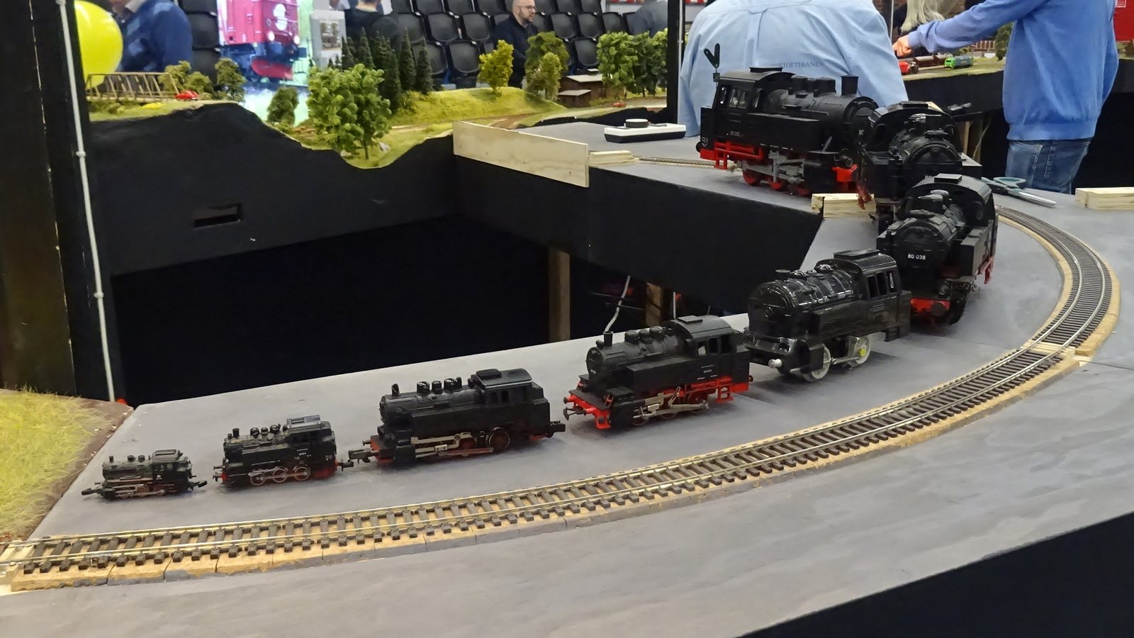 Une même locomotive à vapeur reproduite à plusieurs échelles, de la plus petite à la plus grande, alignée le long d'une voie d'exposition