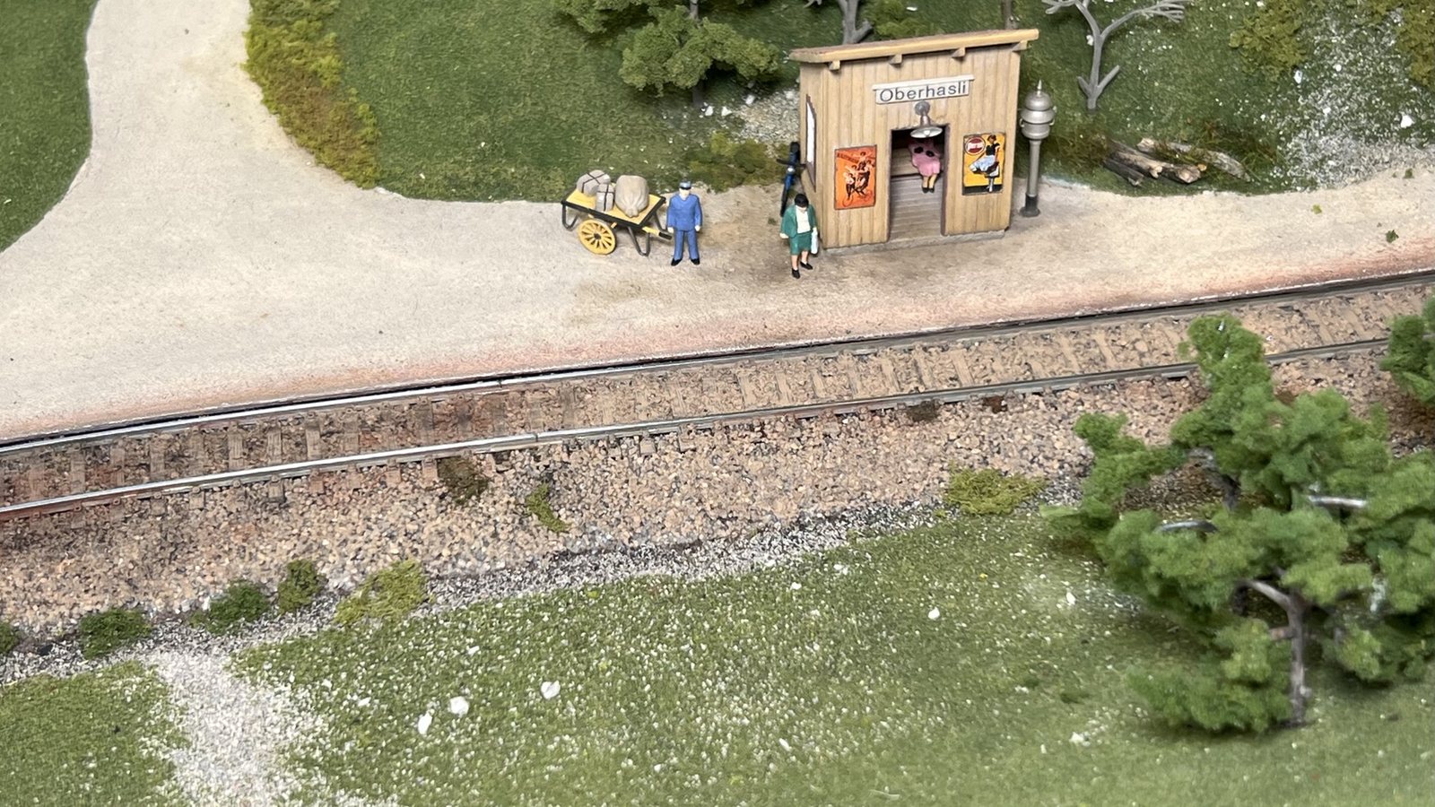 Réseau ferroviaire miniature à l'échelle HO : halte en bois, quai gravillonné, personnages et chariot à bagages le long d'une voie ballastée