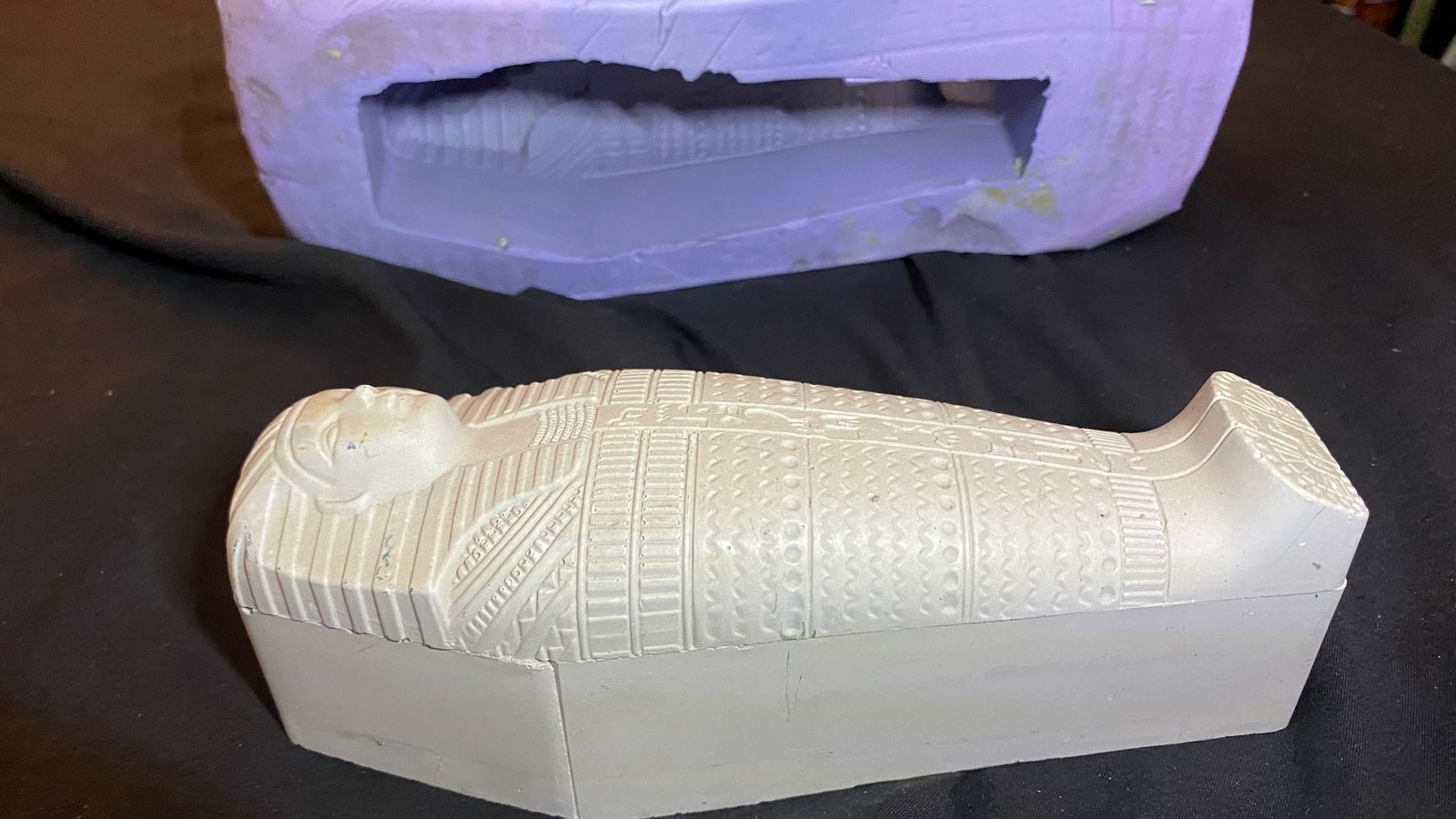 Moule en silicone violet ouvert, posé derrière le tirage en plâtre blanc d’un sarcophage égyptien miniature aux reliefs fins