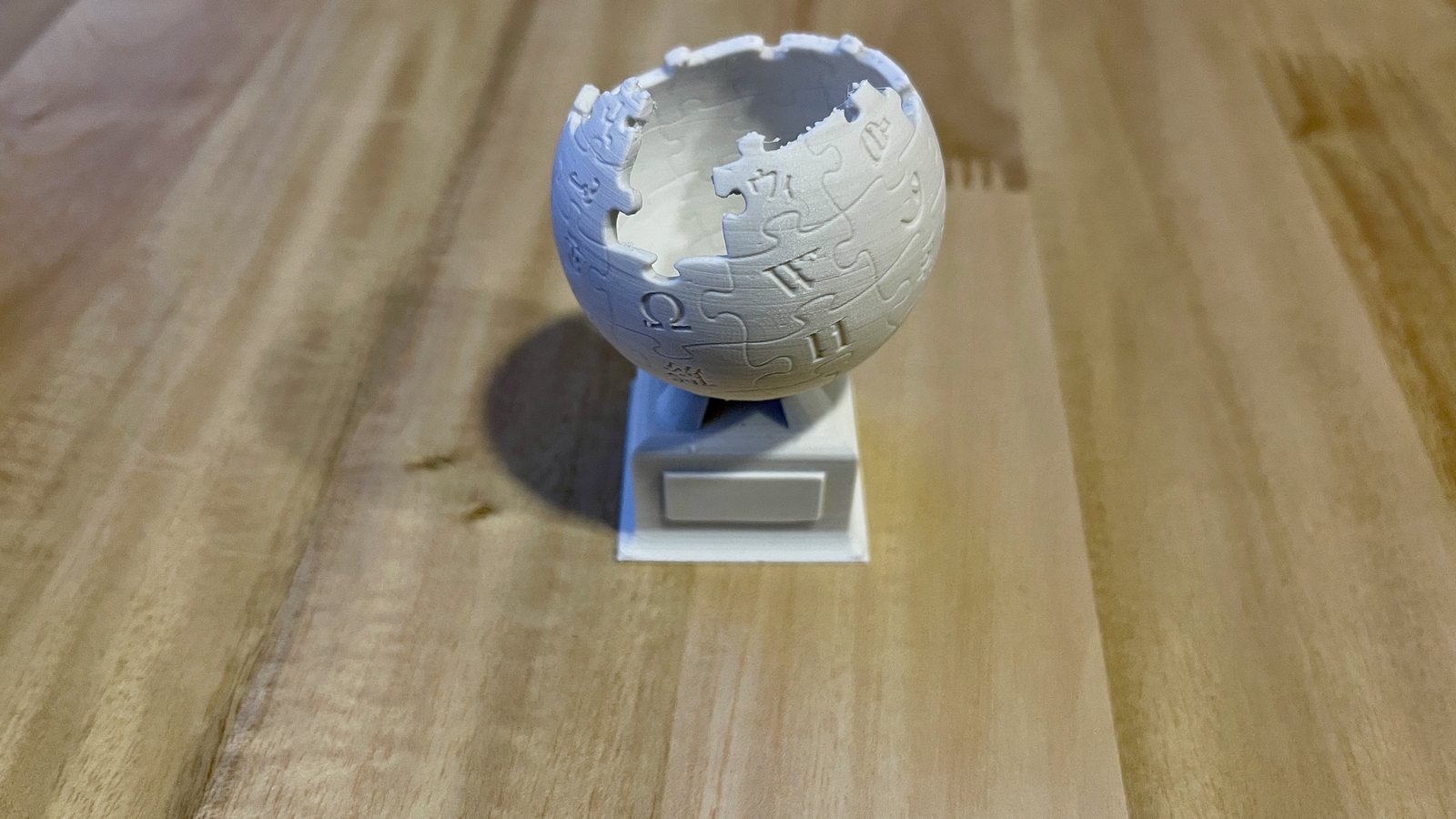 Petit trophée blanc imprimé en 3D en PLA, globe en puzzle sur un socle à plaque, posé sur une table en bois